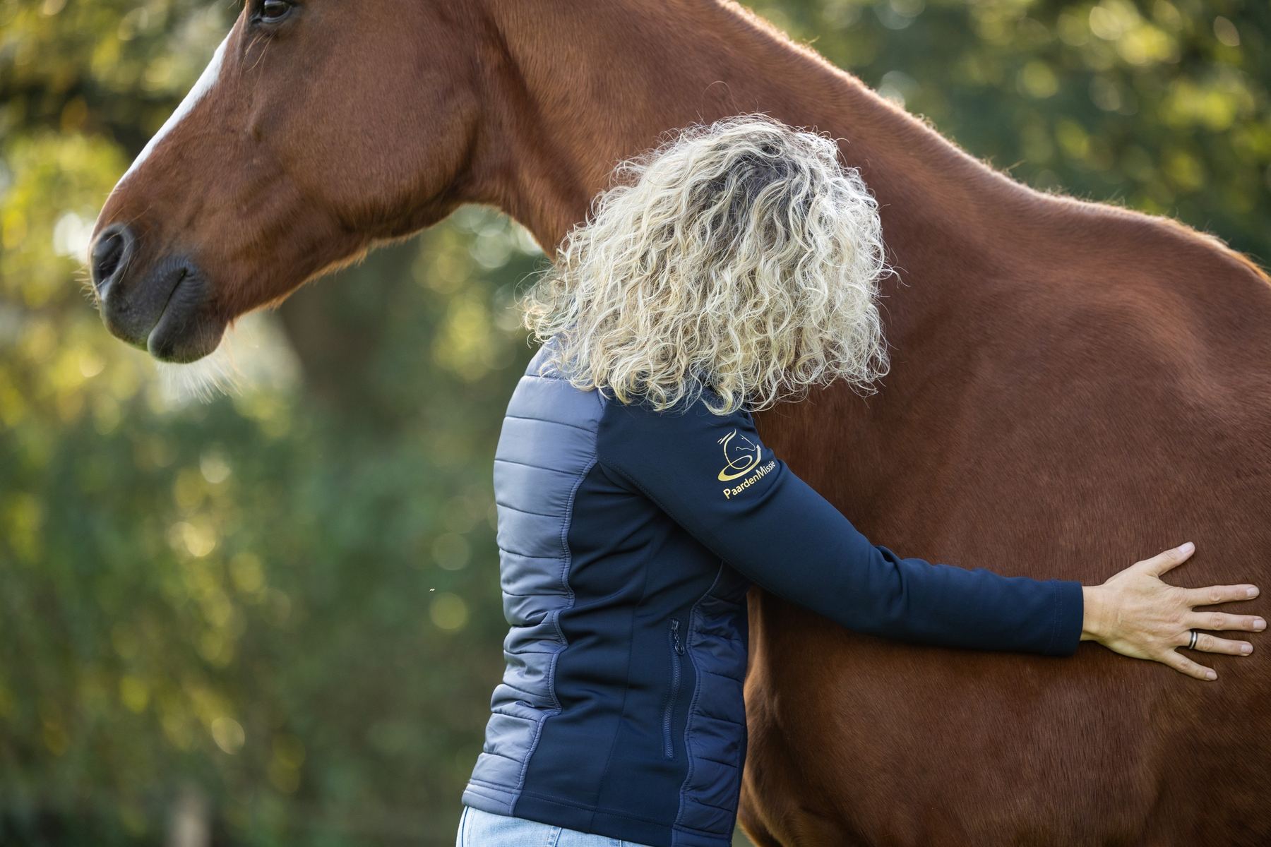 Een vrouw met blonde krullen staat tegen Gorgeous aan en legt haar hand op de flank.