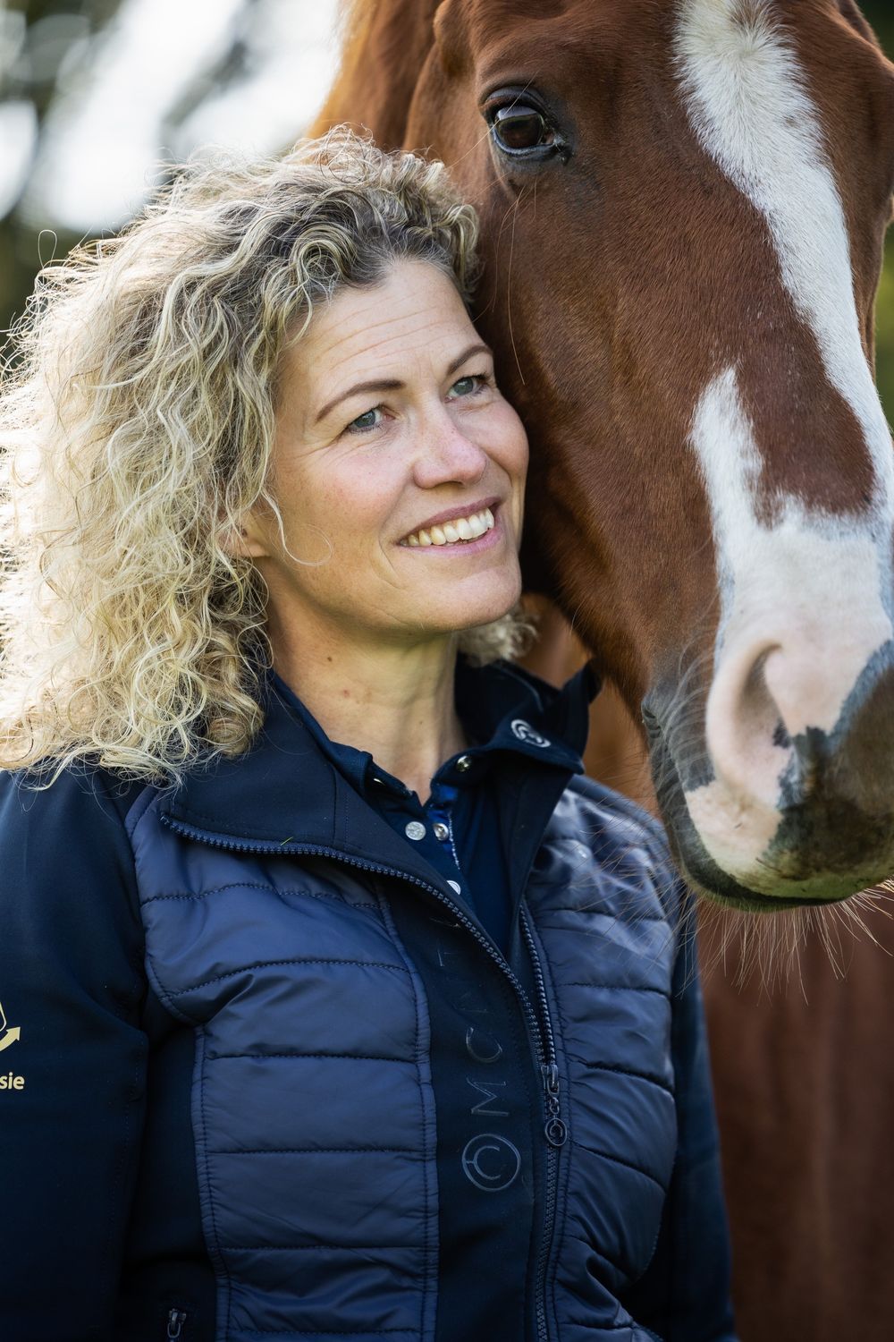 Een vrouw met blonde krullen staat glimlachend dicht tegen het hoofd van Gorgeous.