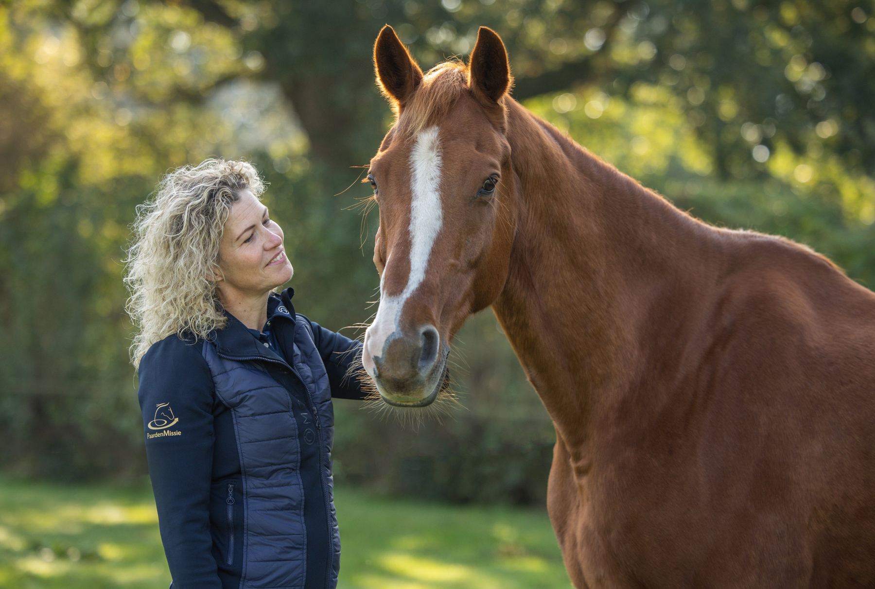 Een vrouw met blonde krullen staat naast Gorgeous en legt haar hand op de wang van het paard.